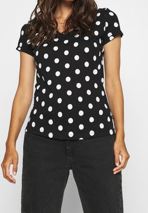 Vrouw draagt een zwart V-hals shirt met korte mouwen en witte polka dots en zwarte broek, zichtbaar van schouders tot heupen.