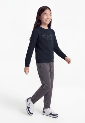 Jeune fille marchant, vêtue d'un sweat-shirt noir EA7, d'un pantalon de survêtement gris et de baskets blanches avec des accents bleu marine sur un fond blanc.