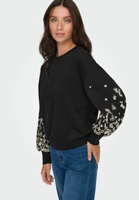 Črna kapuca z dolgimi, napihnjenimi rokavi, okrašena z cvetličnim vezenjem. Okrogla ovratnica in rebrasti manšeti. V kombinaciji z modrimi kavbojkami.