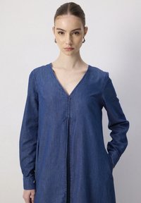Robe en denim avec un col en V, des manches longues et un détail de couture central. Présente une coupe ample et des poches latérales, dans une teinte de bleu moyen.