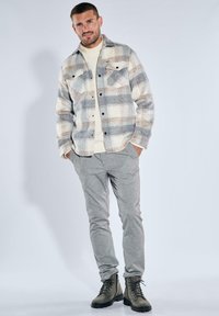 Karierte Flanellhemden in Creme, Grau und Beige mit zwei Brusttaschen; darunter ein cremefarbener Strickpullover; graue schmal geschnittene Hosen; grüne Stiefel.