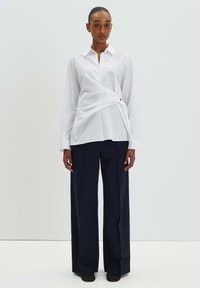 Weißes Langarmhemd mit Wickeldesign, ausgestattet mit einem spitzen Kragen und einer vorderen Drehung. Kombiniert mit weit geschnittenen, marineblauen Hosen.