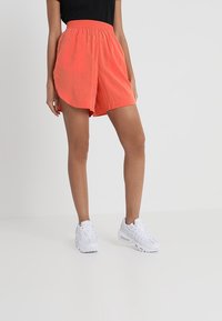 Shorts légers orange avec une taille élastique, présentant un ourlet courbé et un design en couches. Associés à des baskets blanches.