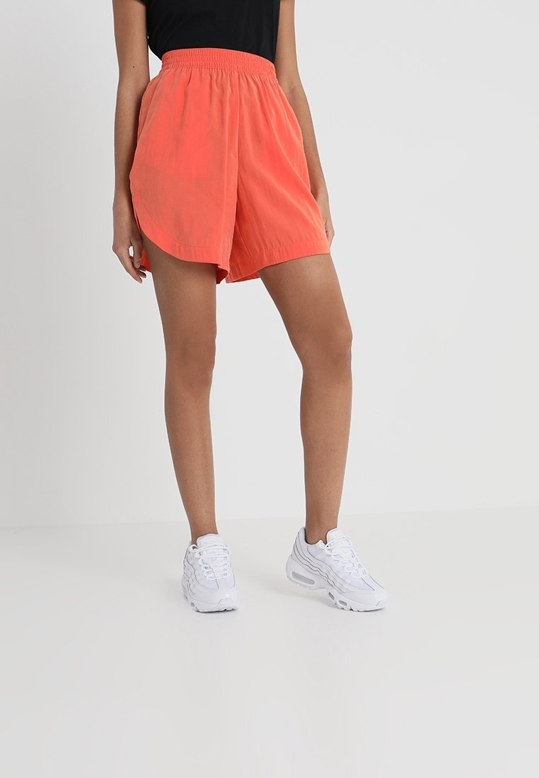 Shorts légers orange avec une taille élastique, présentant un ourlet courbé et un design en couches. Associés à des baskets blanches.