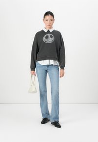 Donna che indossa un felpa nera con simbolo della pace sopra una maglietta a righe bianca, jeans azzurri chiari, scarpe nere, mentre tiene in mano una piccola borsa bianca, su uno sfondo bianco.
