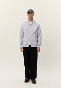 Muž oblečený v svetlosivej mikine na zips, čiernych nohaviciach, tmavohnedých topánkach a tmavosivej šiltovke, stojaci pred jednoduchým svetlým pozadím.