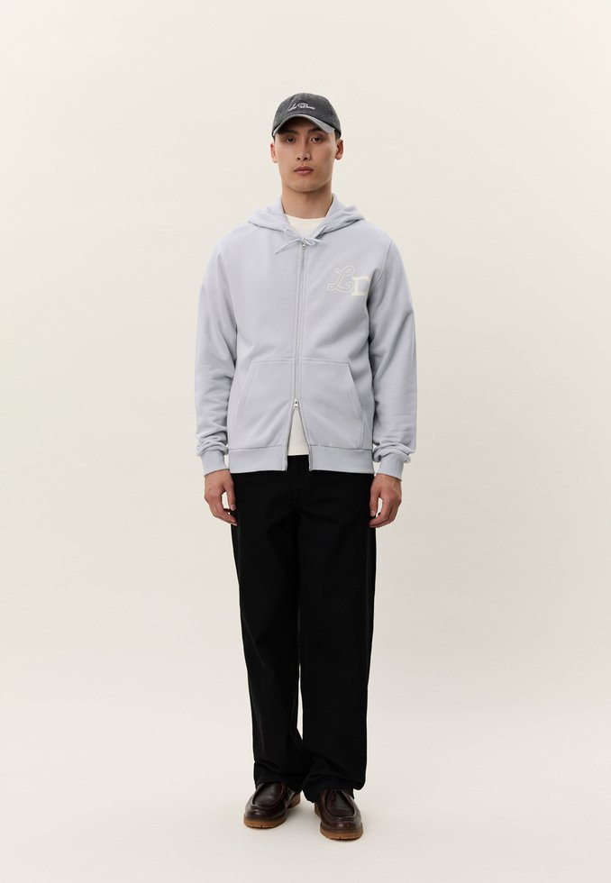 Muž oblečený v svetlosivej mikine na zips, čiernych nohaviciach, tmavohnedých topánkach a tmavosivej šiltovke, stojaci pred jednoduchým svetlým pozadím.
