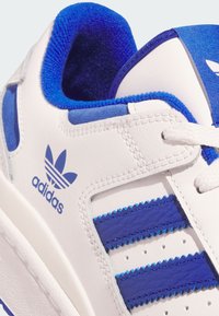 Biele a modré tenisky Adidas zblízka, ukazujúce logo, prešívaný dizajn, biele šnúrky, vypchávkovanú modrú vnútornú podšívku a perforované bočné pruhy.