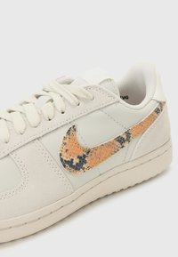 Nike superge z zgornjim delom iz svetlo sive usnje in semškega materiala. Opremljene so z teksturiranim, oranžnim in črnim vzorcem kače na logotipu Swoosh.
