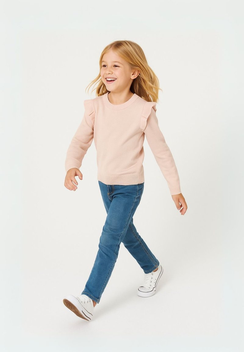 Maglione rosa chiaro a maglia con spalle arricciate, abbinato a jeans in denim blu medio e sneakers bianche. Modella che cammina su uno sfondo neutro.