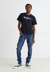 Zwart katoenen t-shirt met witte "Pepe Jeans" logo, gecombineerd met lichtblauwe spijkerbroek en groene sneakers met witte zolen.