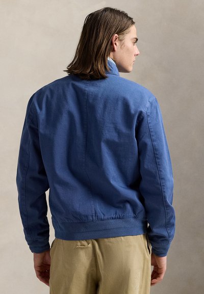 Hombre con cabello castaño hasta los hombros, que lleva una chaqueta azul y pantalones beige, de espaldas sobre un fondo neutro.