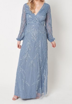 Femme portant une robe bleue longue jusqu'au sol, à manches longues, avec des motifs floraux en sequins argentés et des manches transparentes, associée à des sandales argentées.