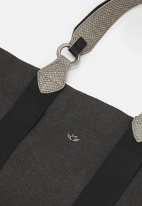 Zwarte stoffen draagtas met een gestructureerde grijze handgreep en een ronde metalen ring. Voorzien van een klein logo met vleugels aan de voorkant.