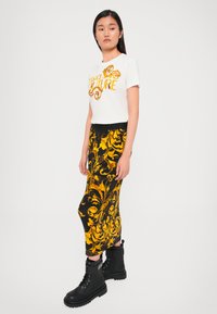 Valkoinen t-paita, jossa on keltainen "Versace Jeans Couture" -teksti, yhdistetty mustaan hameeseen, jossa on keltainen kukkakuosi; mustat nauhakengät paksuilla pohjilla.
