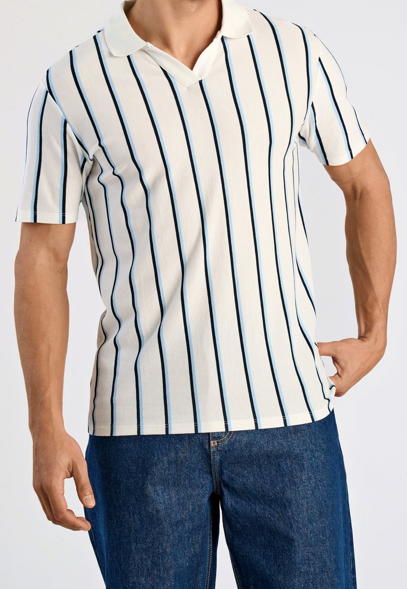 Homme portant un polo blanc à manches courtes avec des rayures verticales noires et bleues, associé à un jean bleu, debout avec une main sur la hanche.