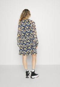 Vestido floral em azul-marinho com flores amarelas e brancas, mangas longas transparentes e cintura franzida. Usado com sapatilhas de cano alto pretas e meias brancas.