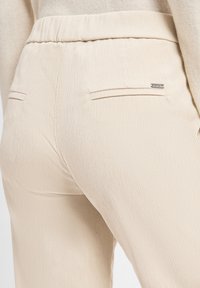 Beige gerippte Hose mit weichem Stoff, elastischem Bund und zwei Gesäßtaschen. Kleines Markenlabel an der rechten Tasche.