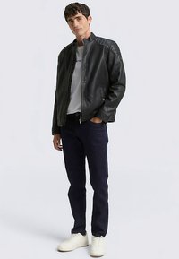 Blouson aviateur en cuir noir avec des accents matelassés aux épaules, porté sur un t-shirt blanc et un jean bleu foncé, assorti à des baskets crème.