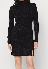 Schwarzes gestricktes Kleid mit langen Ärmeln, Stehkragen, gerippter Textur und seitlichem Schlitz. Der Stoff hat dezente Schimmerakzente.