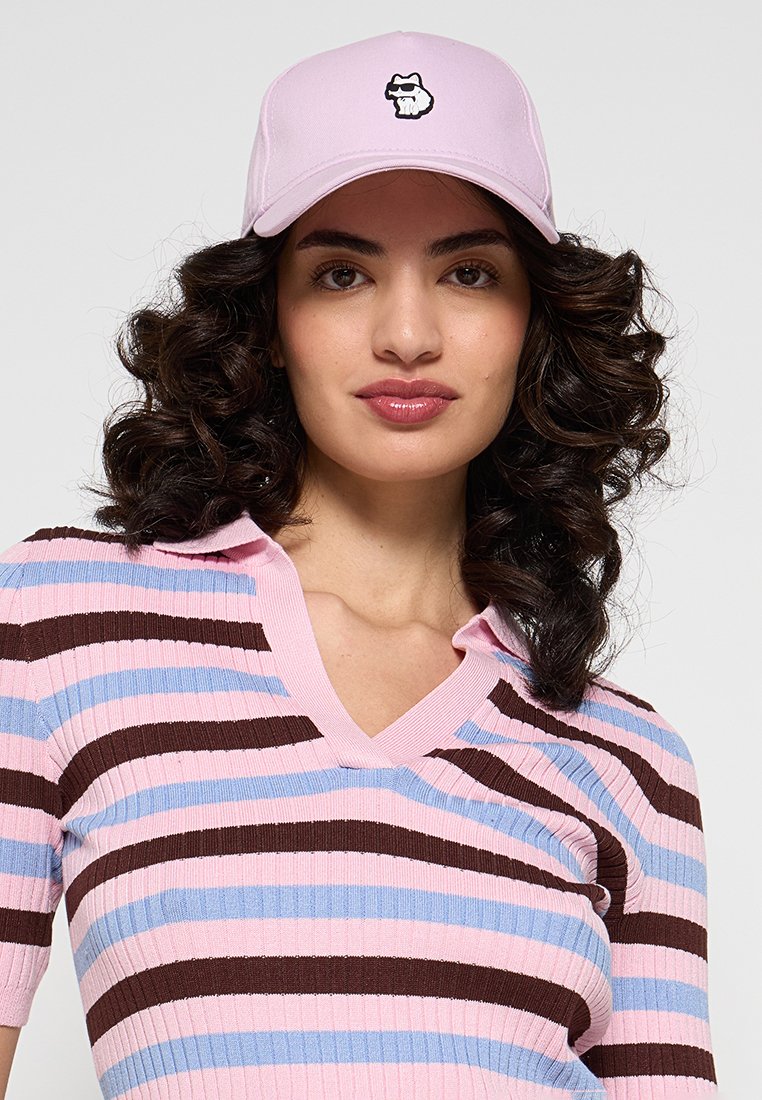 Helle rosa Kappe mit einem gestickten Cartoon-Gesichtlogo; getragen mit einem gestreiften, rippgestrickten Oberteil in Pink, Blau und Braun mit einem V-Ausschnitt.