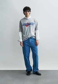 Gray long-sleeve t-shirt layered over a white collared shirt, blue wide-leg jeans, and black shoes, with dark sunglasses accentuating the outfit.