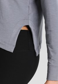 Maglia grigia a maniche lunghe con orlo curvo, caratterizzata da uno spacco laterale, abbinata a leggings neri. Il materiale sembra morbido e leggermente testurizzato.