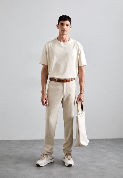 Man wearing beige t-shirt, matching pants, brown belt, and sneakers, holding a cream tote bag, standing against a plain light gray wall.