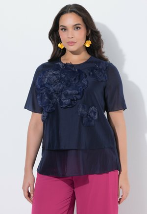 Kobieta w granatowej bluzce z dużymi aplikacjami kwiatowymi, żółtymi kolczykami w kształcie kwiatów i jasnoróżowych spodniach, stojąca na białym tle.