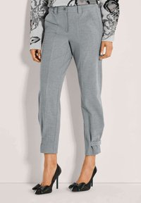 Vrouw in lichtgrijze, op maat gemaakte enkellange broek met omslagbandjes, gecombineerd met zwarte hoge hakken met strikdetail.