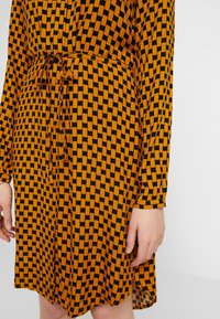Robe à manches longues avec un motif à carreaux noir et jaune moutarde, boutonnée sur le devant et ceinture nouée à la taille, montrée du milieu de la cuisse au torse.