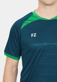 Mörk tealfärgad sporttröja med gröna detaljer på kragen och ärmarna. Har en subtil struktur och bokstaven "FZ" tryckt i vitt.