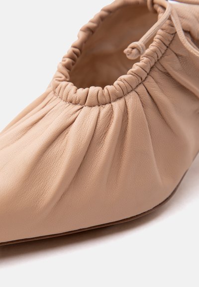 Ευλύγιστο μπεζ δερμάτινο μπαλαρίνα παπούτσι με ραμμένη ελαστική περίμετρο και μικρό φιόγκο δεμένο στο άνοιγμα, εμφανίζεται σε κοντινή λήψη.