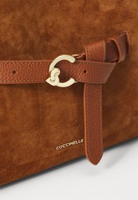Brown suede handbag with a textured brown leather strap and gold-tone hardware, featuring a distinctive looped metal accent and logo detail.