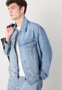 Light blue denim jacket with a classic cut, two front pockets, and a small blue label. Worn over a white shirt, held with a black bag strap.