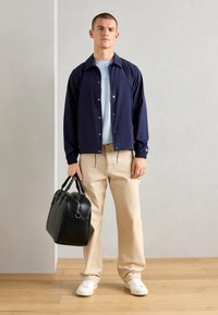 Navy lightweight jacket with snap buttons, paired with a light blue t-shirt. Beige trousers and white sneakers. Holding a black leather bag.