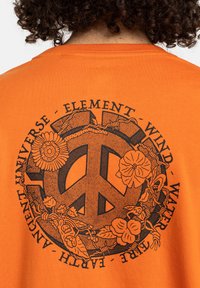 Camiseta de algodón naranja con un gran gráfico de signo de paz negro rodeado de elementos florales y el texto: "UNIVERSO - ELEMENTO - VIENTO - AGUA - FUEGO - TIERRA."