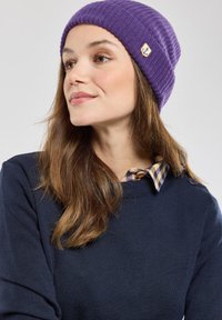 Paarse gebreide beanie met een geribbelde textuur en een klein geel logo标签. Draagt met een marineblauwe trui en een geruite coltrui eronder.