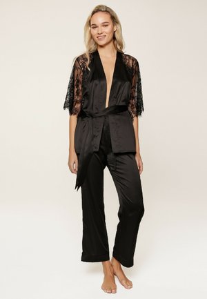 Femme debout pieds nus portant un pyjama en satin noir avec des manches en dentelle aux coudes et une large ceinture à la taille, souriant devant un fond uni.