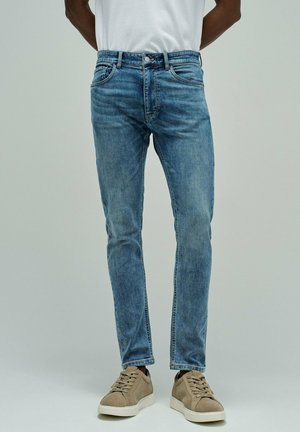 Persona con jeans ajustados azules y zapatillas beige, de pie contra un fondo liso de color gris claro con las manos detrás de la espalda.