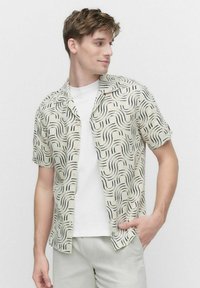 Korte mouwen shirt in lichtbeige met een golvend zwart patroon. Het heeft een kampkraag en een knoopsluiting aan de voorkant, gedragen over een wit T-shirt.
