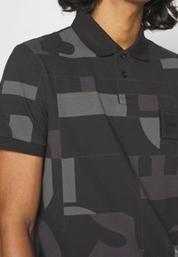 Polo noir avec des motifs géométriques en nuances de gris, doté d'un col, de deux boutons et d'un badge en tissu portant le nom de la marque.