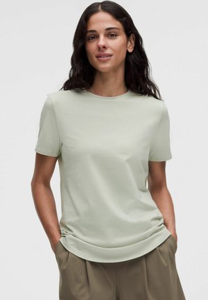 Kobieta z długimi ciemnymi włosami, ubrana w jasnozielony t-shirt z krótkim rękawem i oliwkowe spodnie, stoi z rękami w kieszeniach na jednolitym tle.