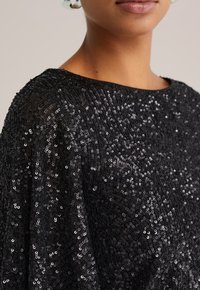 Top nero di paillettes con scollatura ampia, che mostra una texture scintillante e una vestibilità svasata. La vista ravvicinata mette in evidenza i dettagli intricati delle paillettes.