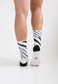 Weiße Socken mit schwarzen diagonalen Streifen, ausgestattet mit einem gerippten Bund und schwarzen Akzenten an Ferse und Zehen. Der Stoff wirkt weich und flexibel.