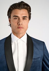 Blå smokingjacka med svarta satinaxlar, texturerad tyg och en vit skjorta under. Fickdetalj på vänster sida.