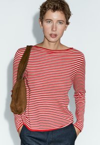 Jongere met kort haar, gekleed in een rood-wit gestreepte lange mouw shirt, donkere broek en met een bruine schoudertas.