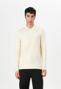 Polo à manches longues en crème clair, fabriqué en doux tissu de coton. Doté d'un patte de boutonnage à trois boutons et d'un petit logo texturé sur la poitrine.