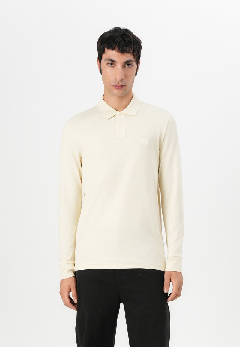 Polo à manches longues en crème clair, fabriqué en doux tissu de coton. Doté d'un patte de boutonnage à trois boutons et d'un petit logo texturé sur la poitrine.