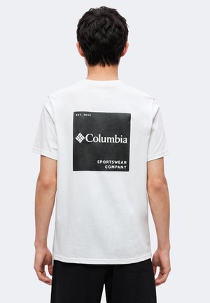 Hombre de espaldas lleva una camiseta blanca con un logo cuadrado negro y texto "Columbia Sportswear Company Est. 1938" en la parte trasera.